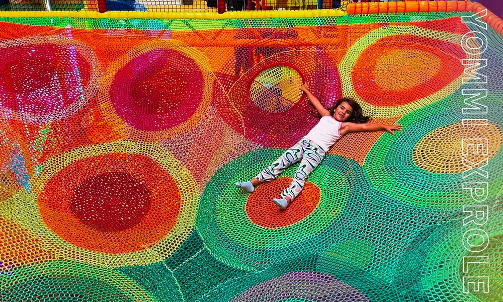 Hand-woven, colorful suspended rainbow net attraction in an indoor playground, maximizing vertical space while providing a fully enclosed safe play area.