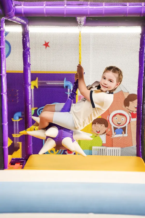 Young girl smiling while riding a suspended purple soft spike swing in the play structure