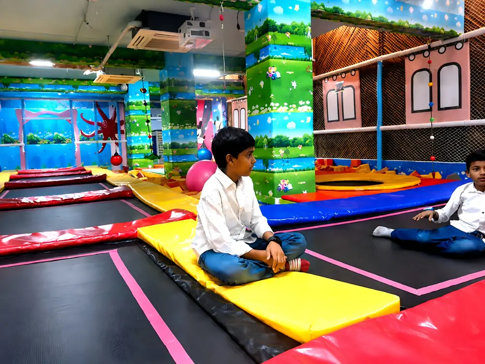 Multi-color padded trampoline zone with safety foam edges inside commercial indoor soft playground, kids resting on trampoline mats